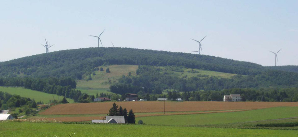 seven wind turbines rising above tree covered mountain behind farms